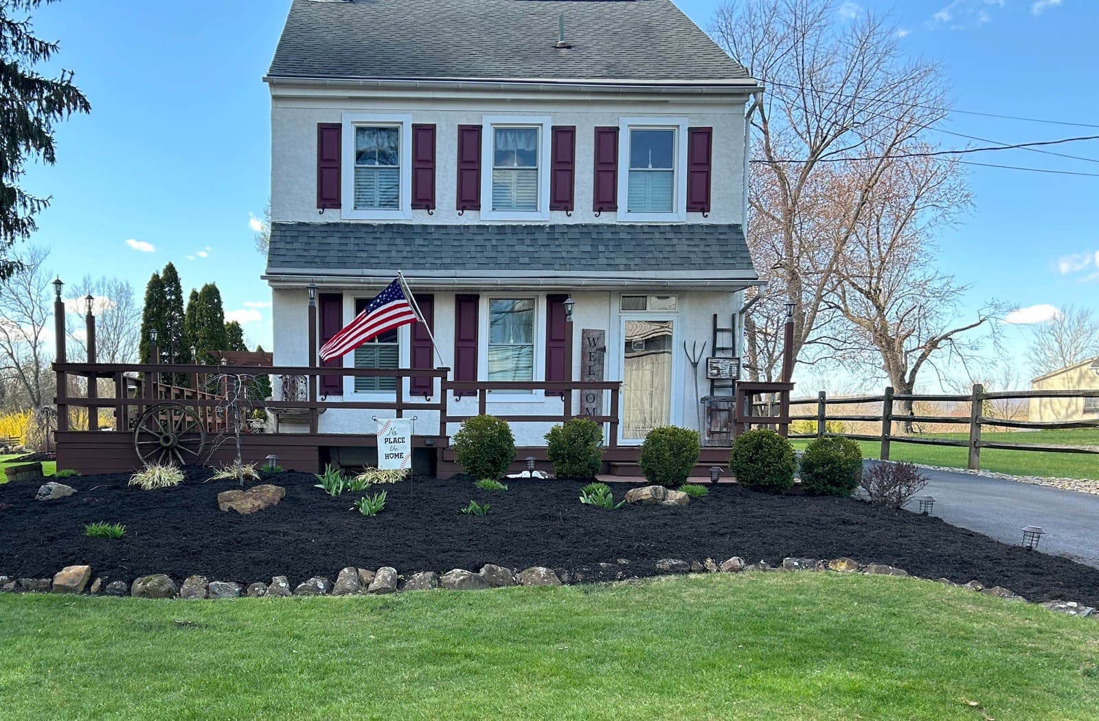 Large stone-bordered front bed at a stucco farmhouse, cleared and freshly mulched around shrubs